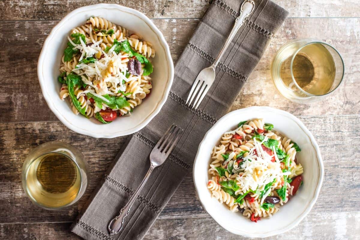 overhead of two bowls of arugula pasta salad on a table with two glasses of wine.