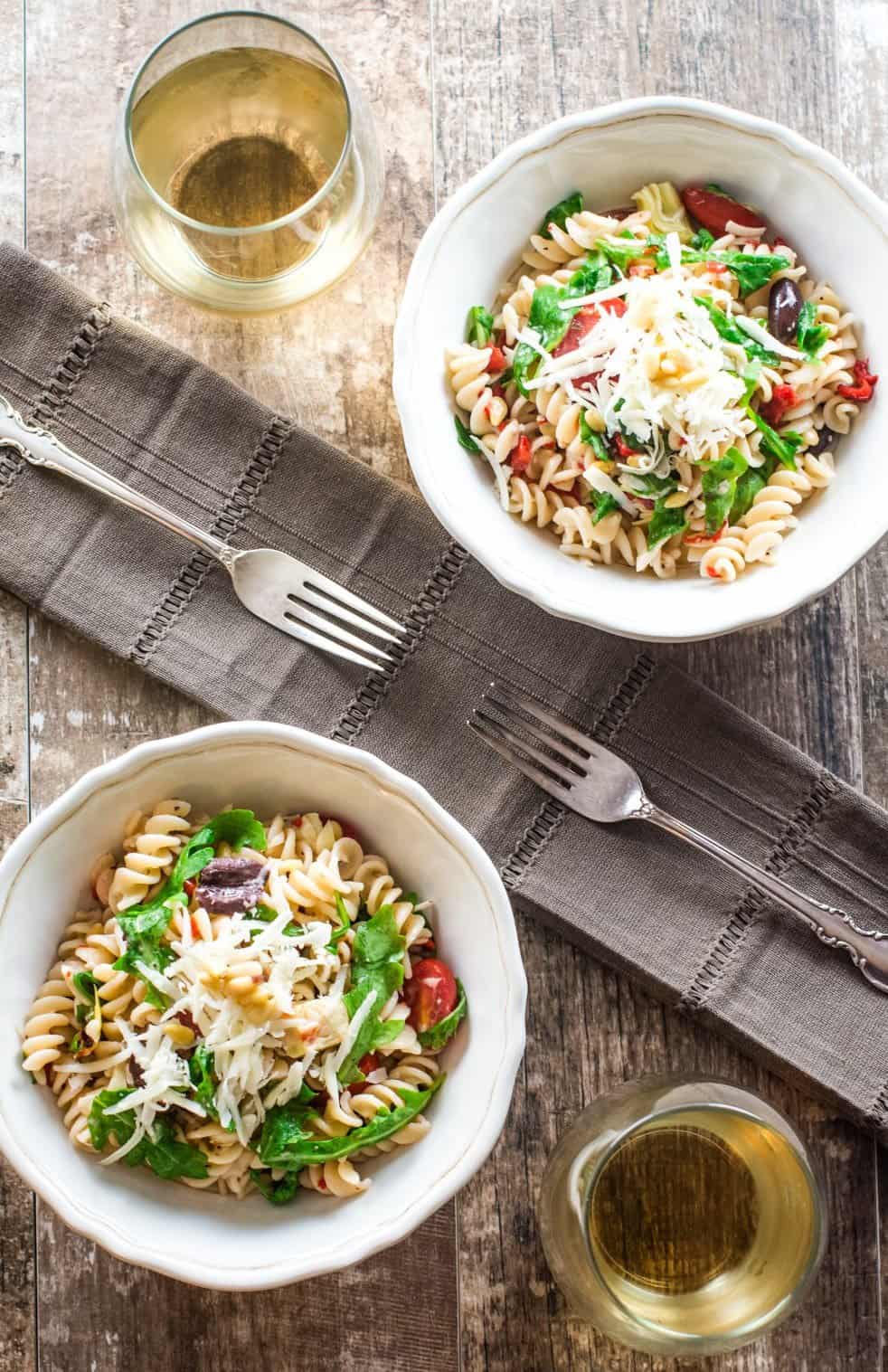 two bowls of pasta salad on a table with two forks and two glasses of wine.