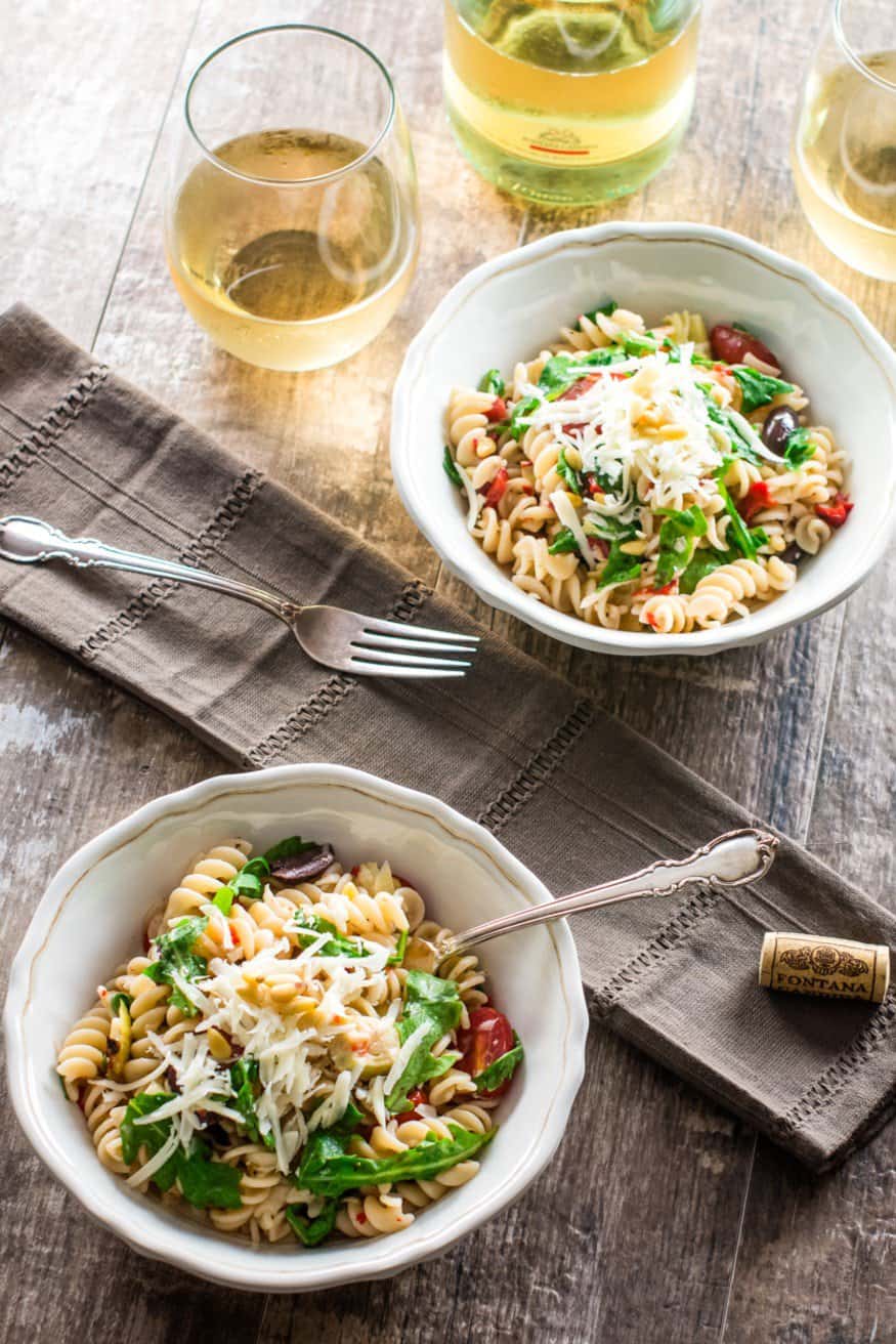 arugula pasta salad in two bowls with a bottle of wine and glasses in the background.