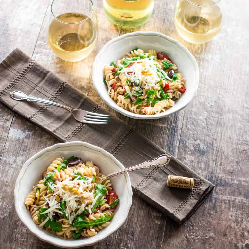 two white bowls with pasta salad and wine in the background.