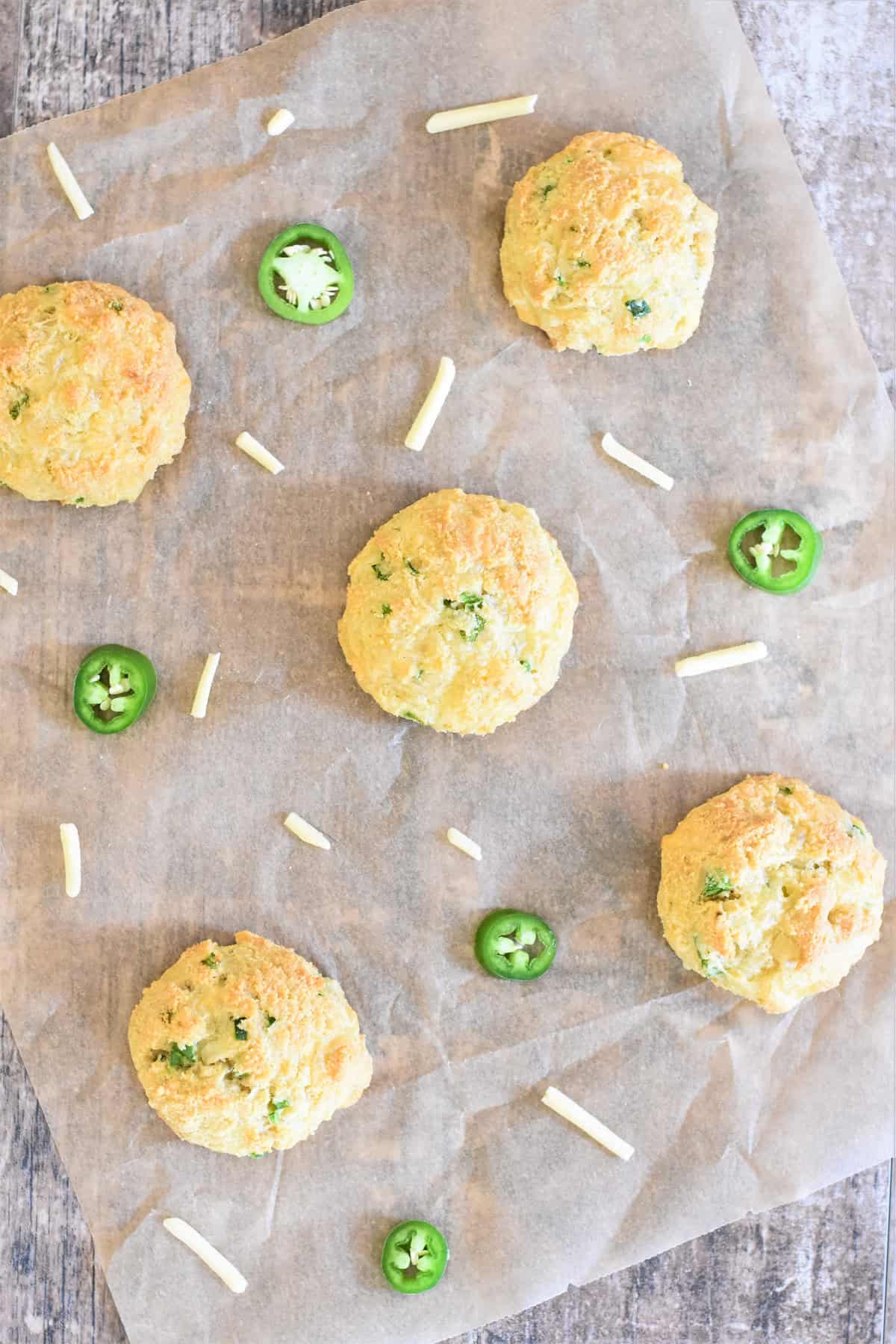 Biscuits on parchment paper with slices of jalapeno and shreds of cheese around them.