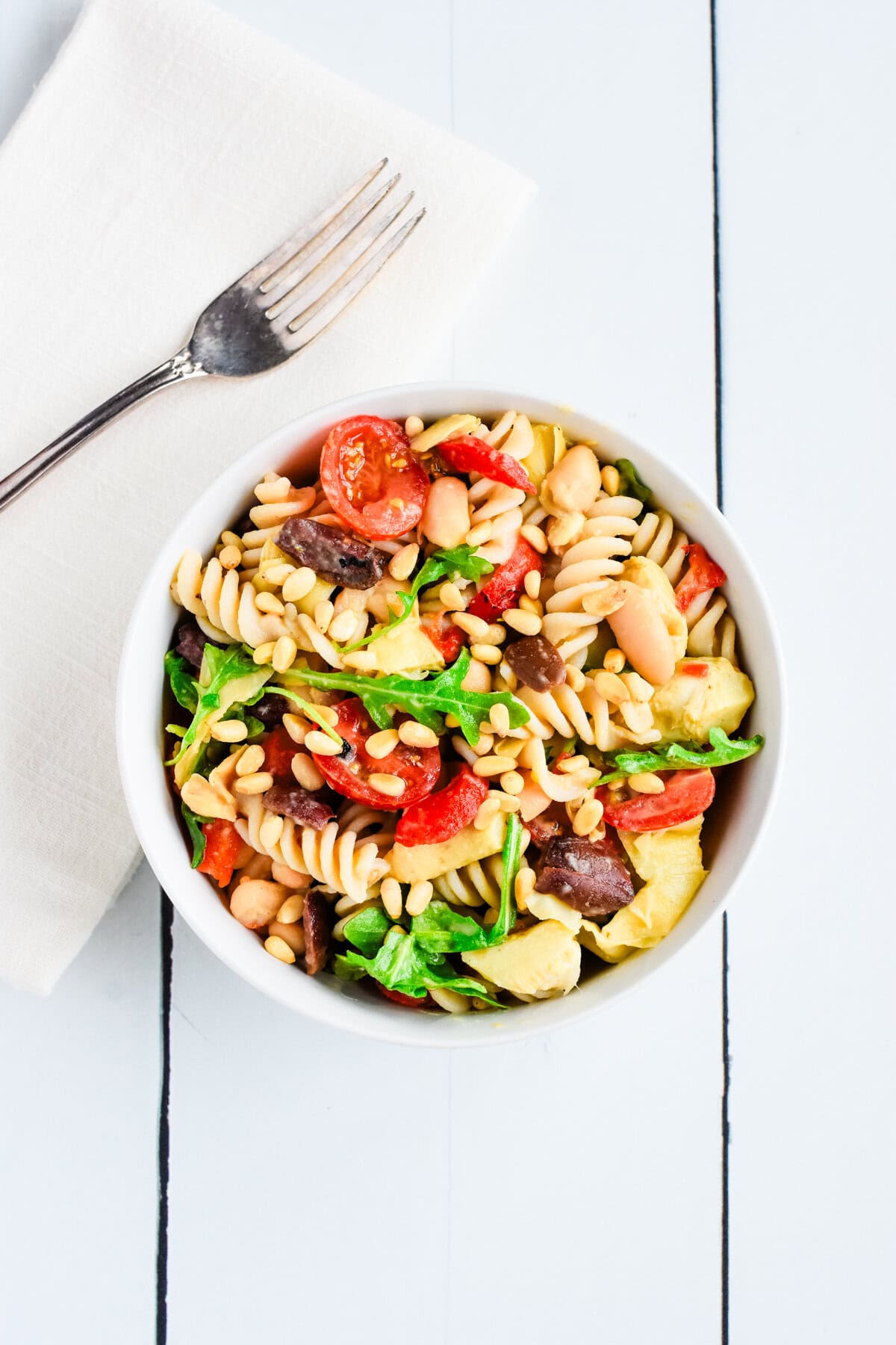 A white bowl of lemony arugula pasta salad with a kitchen napkin on the side and a fork on it.