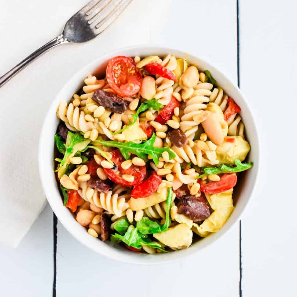 A white bowl filled with a colorful pasta salad and a fork on a kitchen napkin next to it.
