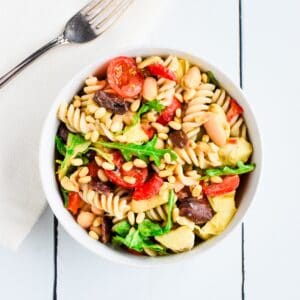 A white bowl filled with a colorful pasta salad and a fork on a kitchen napkin next to it.