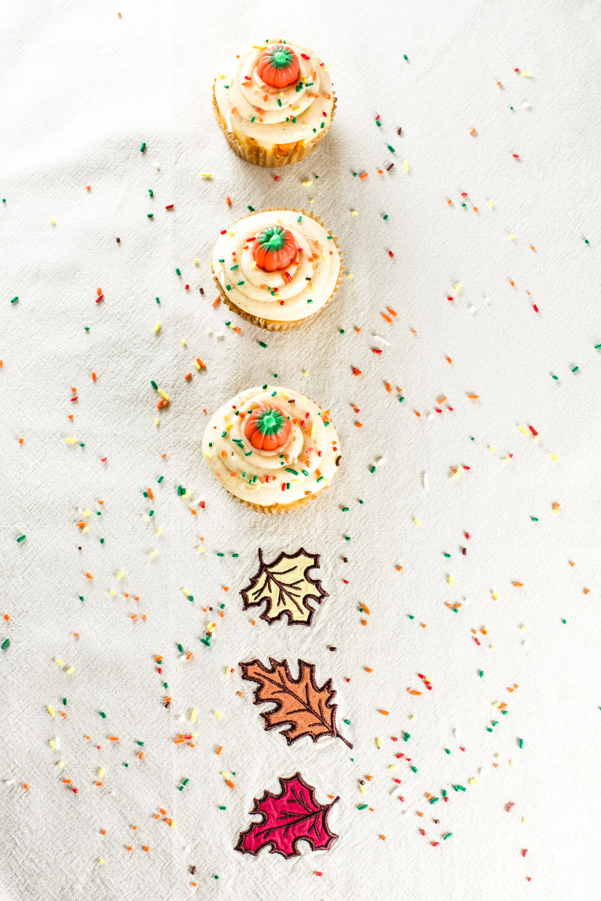 Three cupcakes on a kitchen towel with a leaf design and sprinkles all around.