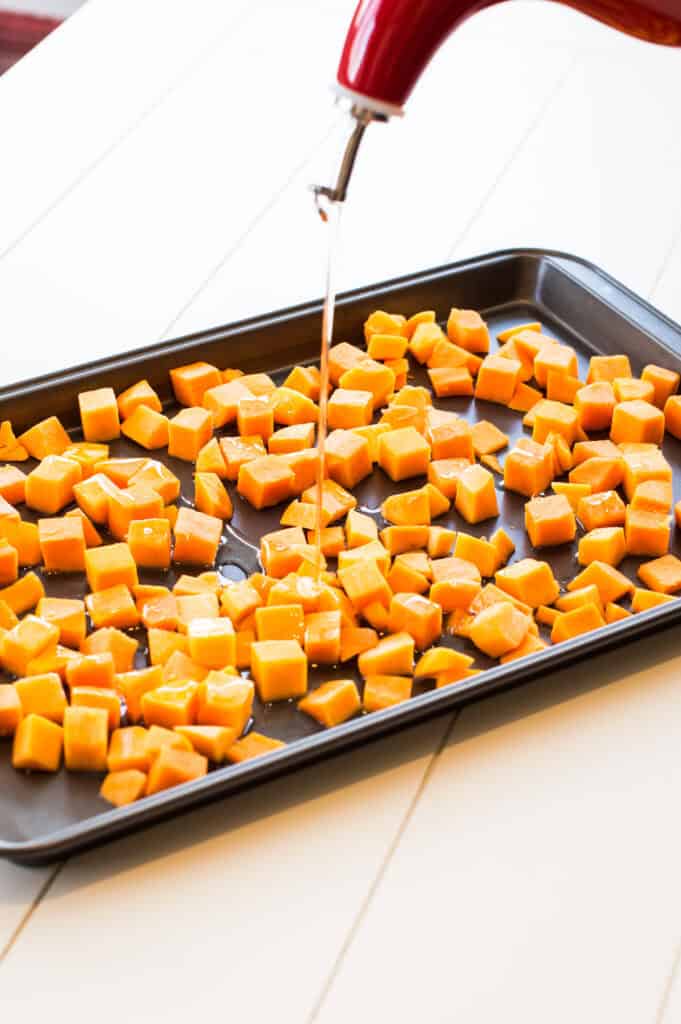 Pouring olive oil onto raw cubes of butternut squash on a baking sheet.