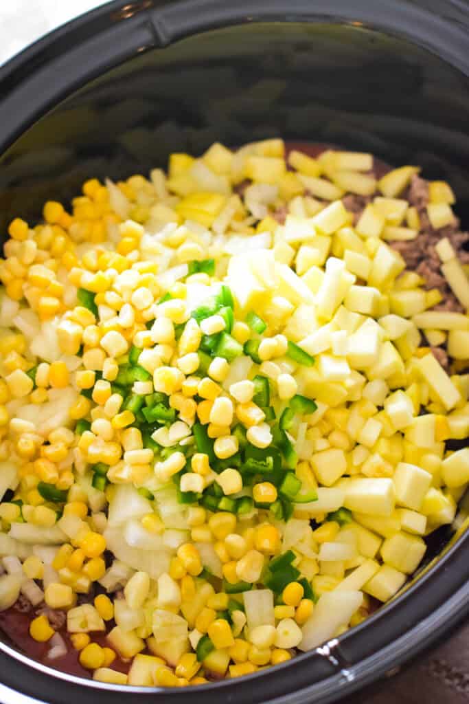 Veggies and spices added to canned tomatoes, ground meat, and beans in a crockpot.