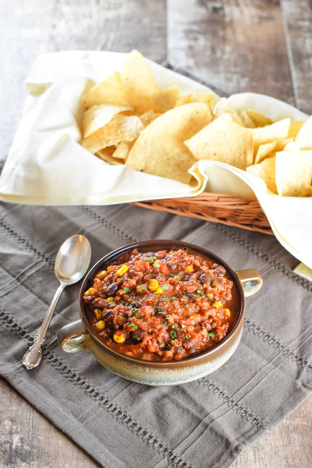 A serving of chili in a bowl with a spoon next to it and a basket of tortilla chips behind it.