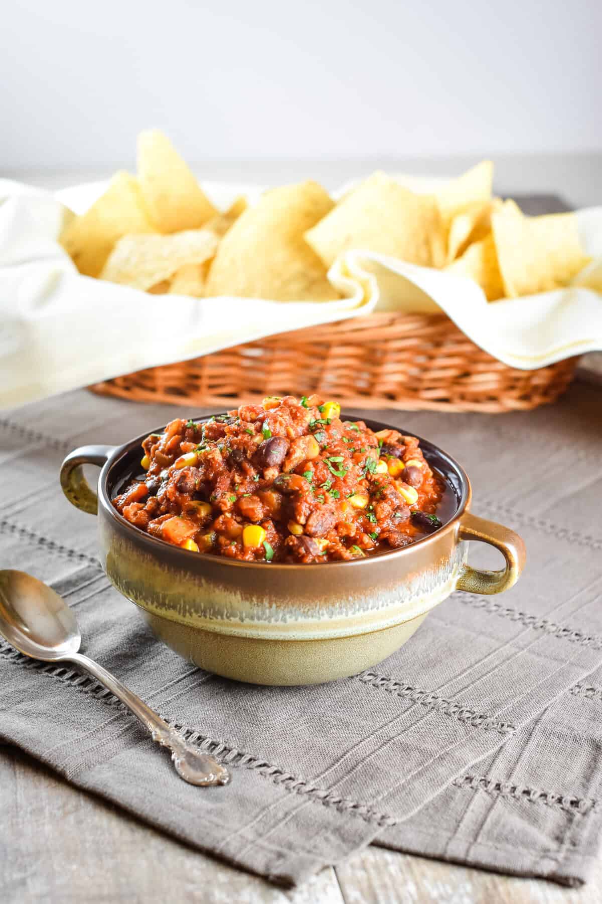 A bowl of chili with pork and beans on a kitchen napkin with a spoon next to it and a basket of tortilla chips in the background.