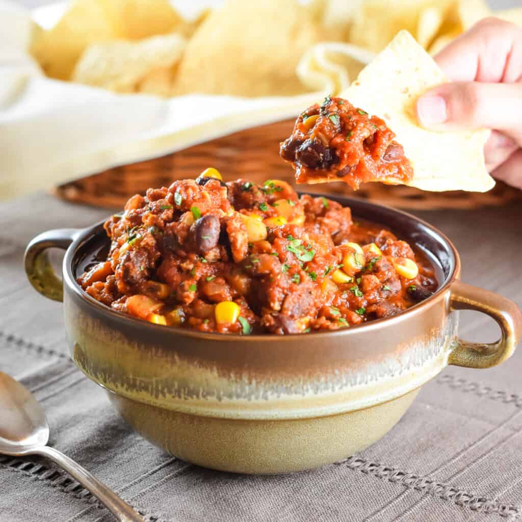 A hand holding a tortilla chip after scooping it into a bowl of chili.