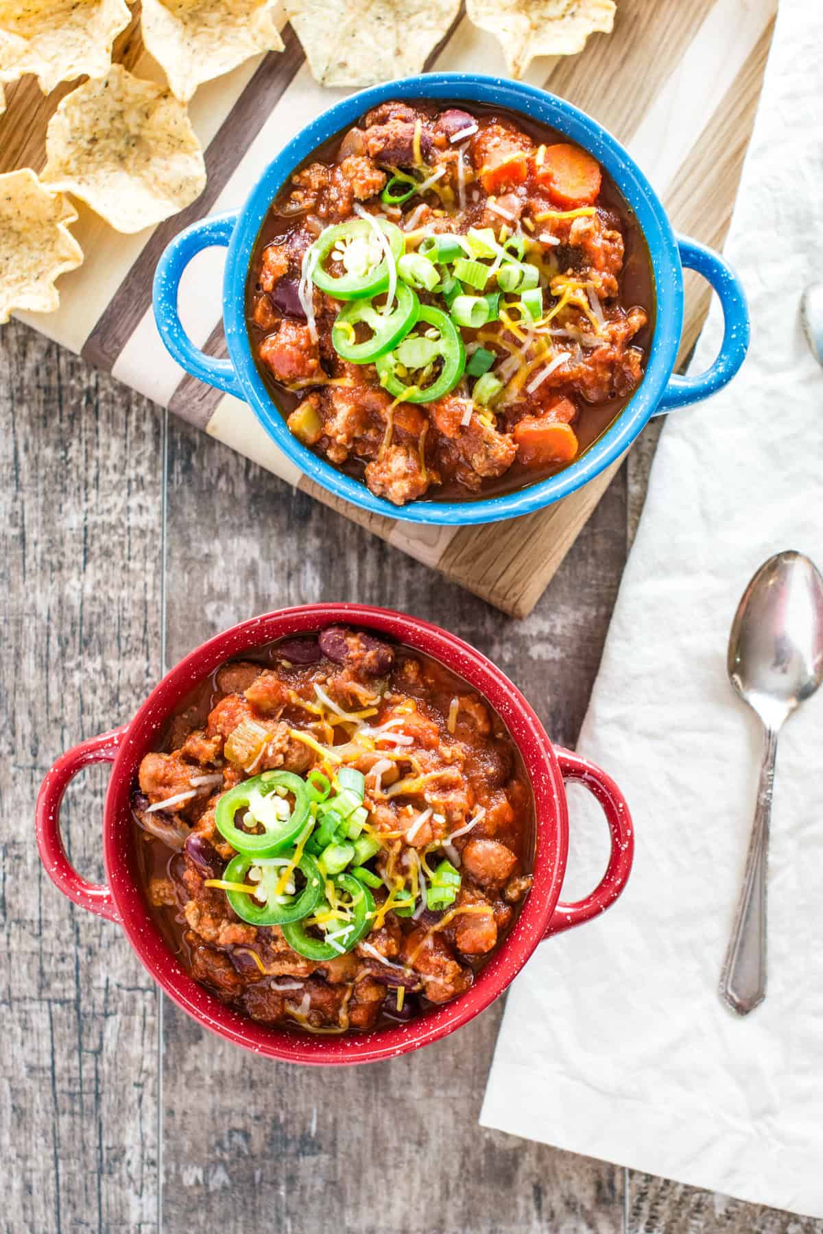 A red bowl and a blue bowl of turkey chili with shredded cheese, scallions, and sliced jalapenos on top.