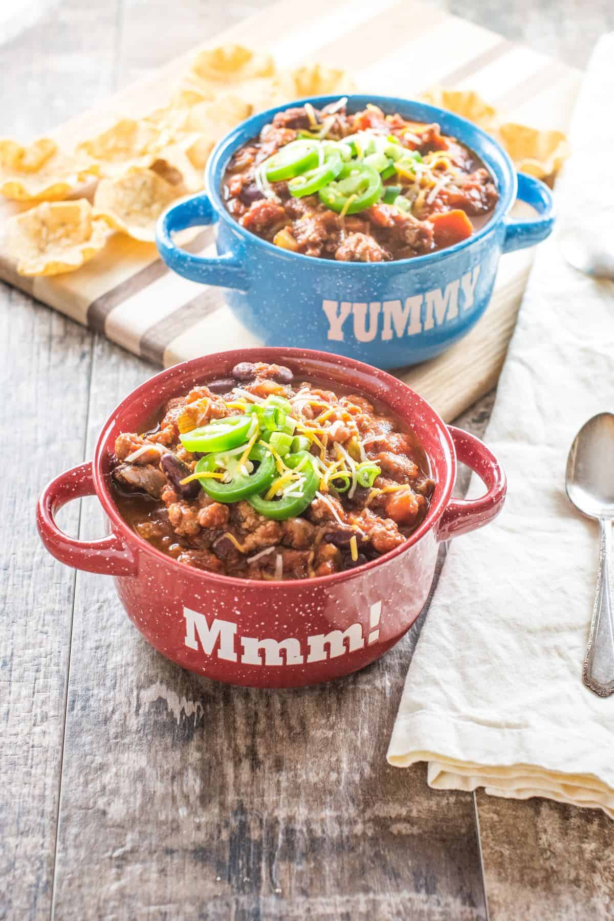 Two bowls of chili with spoons on a kitchen napkin next to them and a wooden board of tortilla chips behind them.