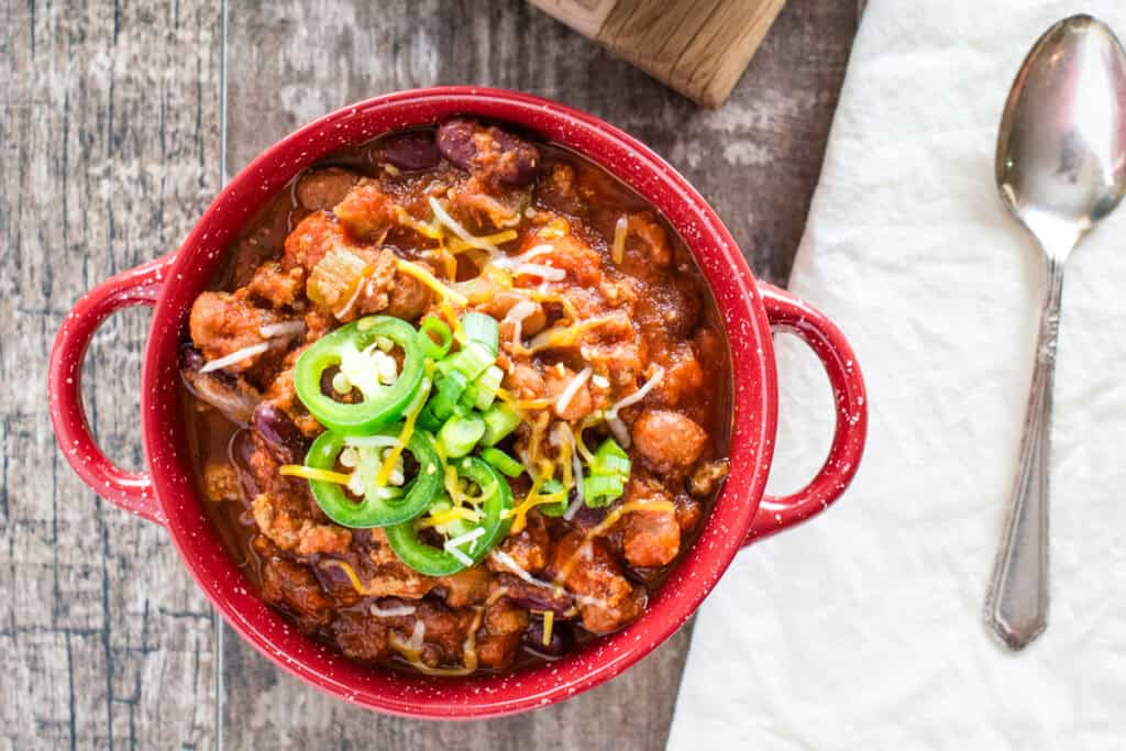 Turkey chili in a red bowl with scallions, shredded cheese, and jalapenos on top and a spoon next to it.