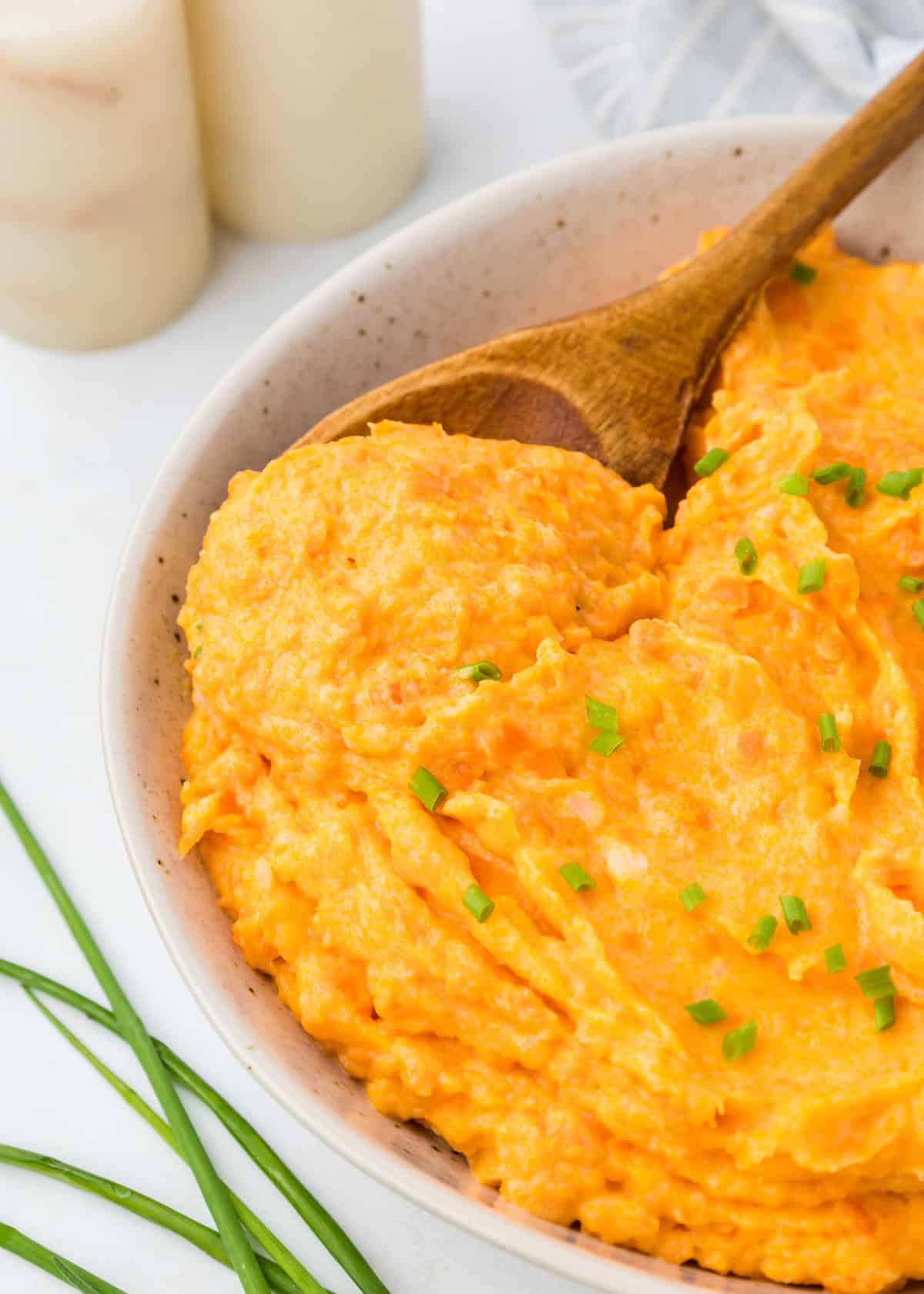 Carrot mashed potatoes in a white dish with a wooden spoon in it.