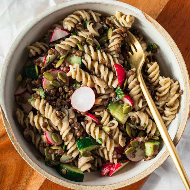 Vegan lentil pasta salad in a bowl with a fork sitting on a wooden board.