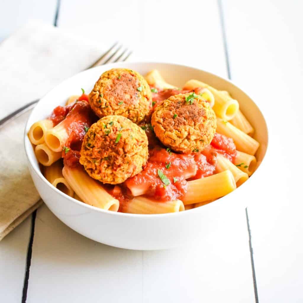 A white bowl of rigatoni pasta with marinara sauce topped with three chickpea balls.