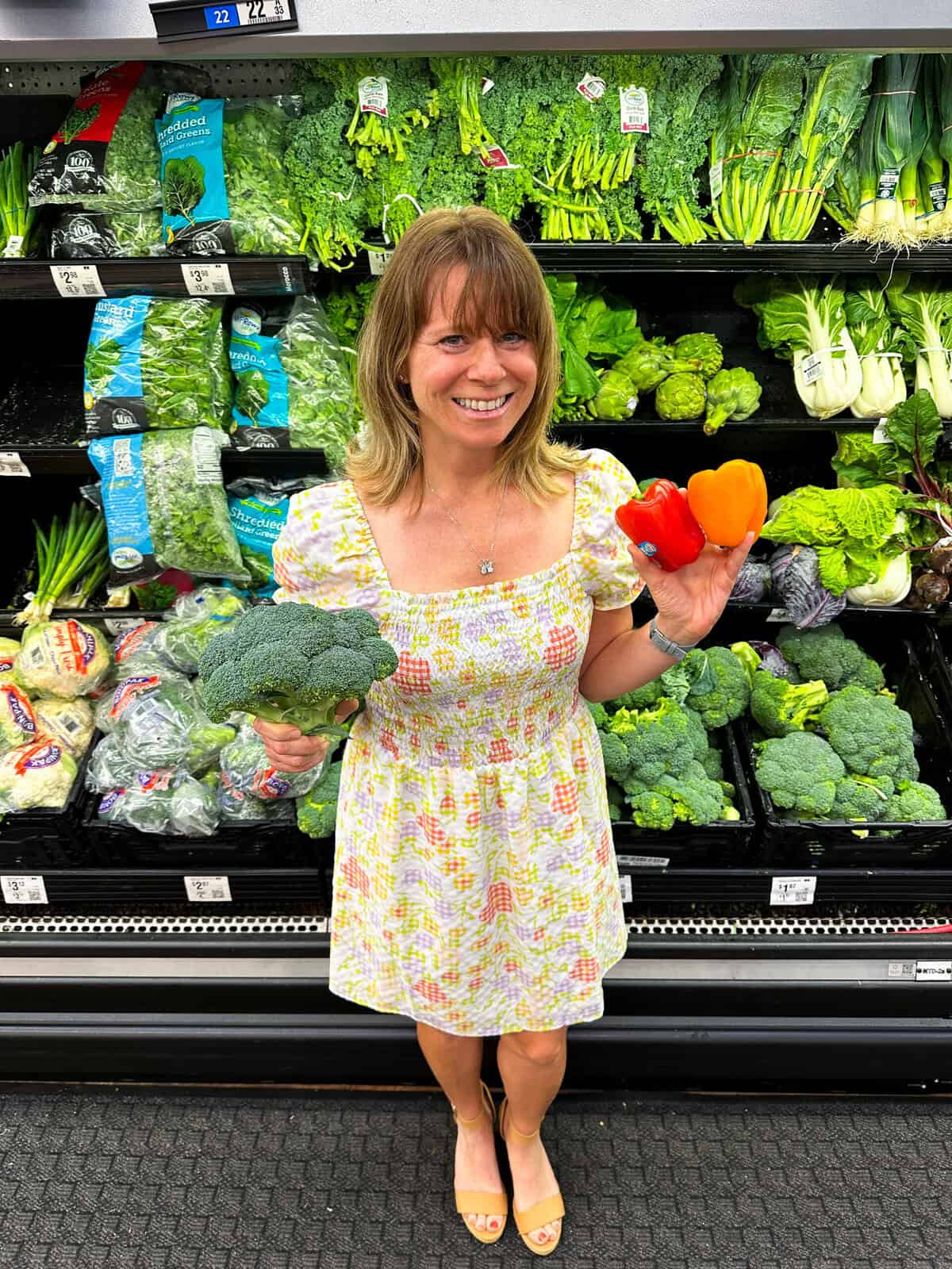 blog author holding broccoli and peppers in front of the produce section at the grocery store.