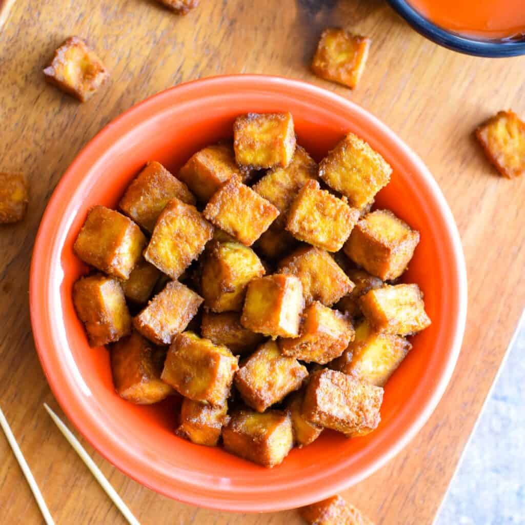 Tofu bites in an orange bowl on a wooden board.