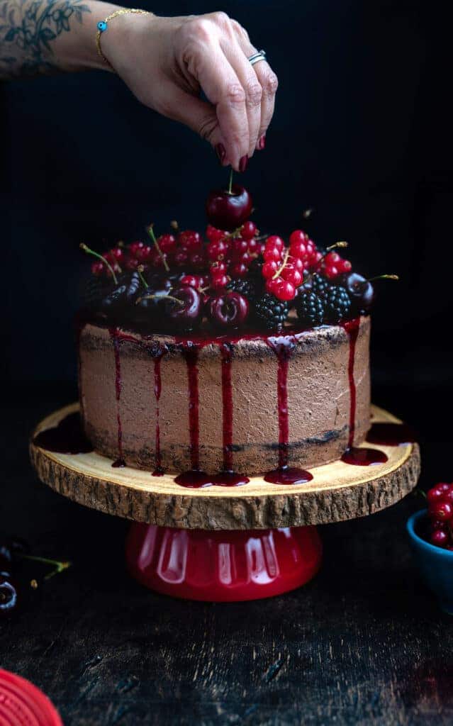 A hand putting a cherry on top of a vegan chocolate cake.