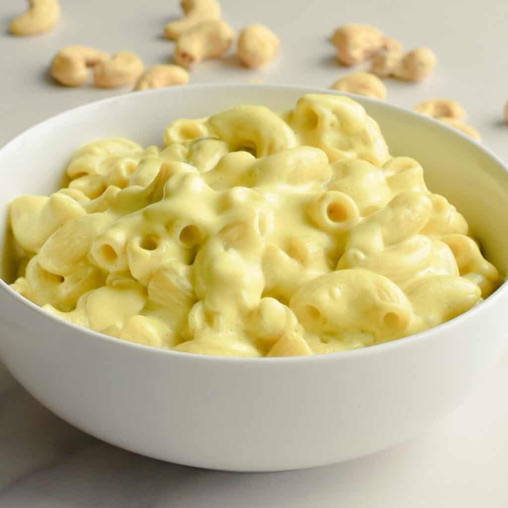 Close-up of cashew mac and cheese in a white bowl with cashews on the table behind it.
