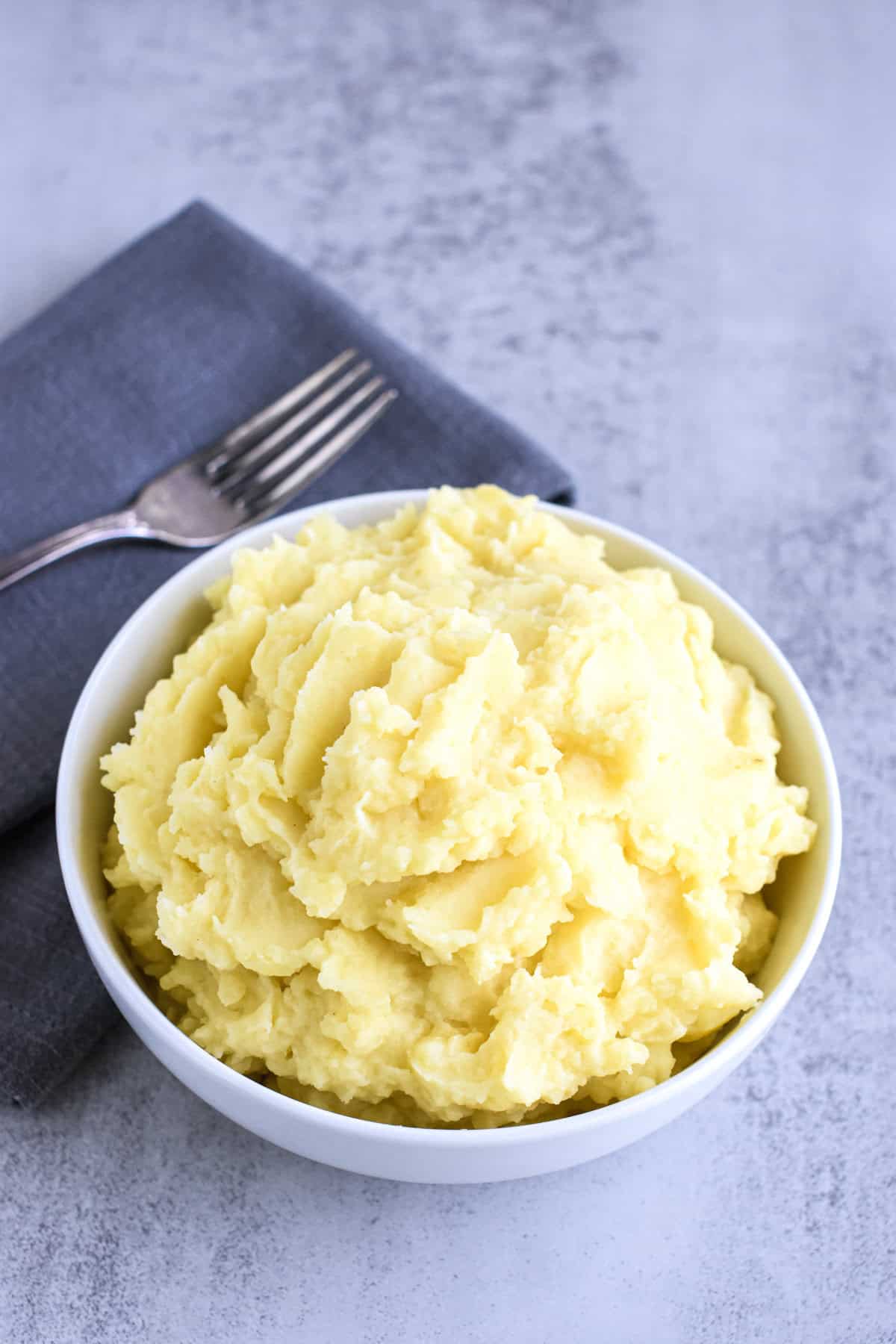 A white bowl with mashed potatoes and a fork on a kitchen napkin next to it.