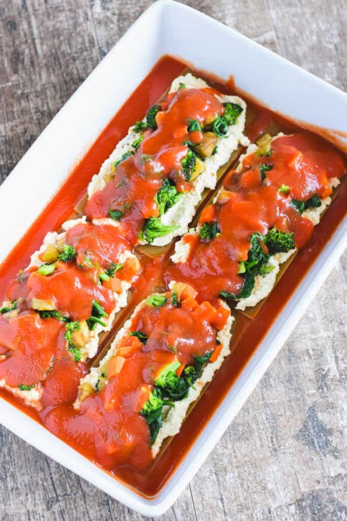 Tomato sauce on top of the first layer of vegan cheese and veggies on lasagna sheets in a white casserole dish.