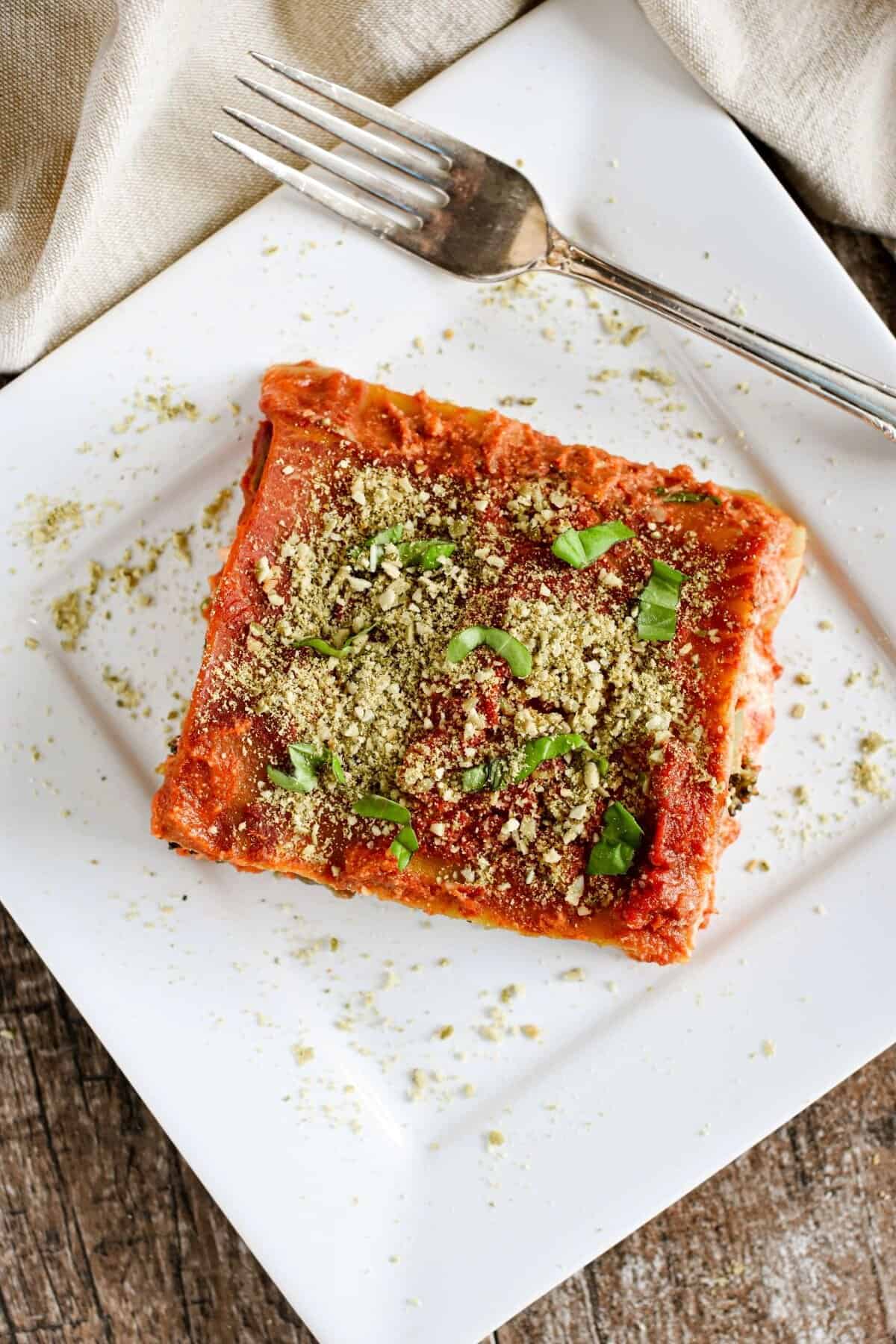 The top of a slice of lasagna garnished with vegan Parmesan and basil on a white plate with a fork on it.