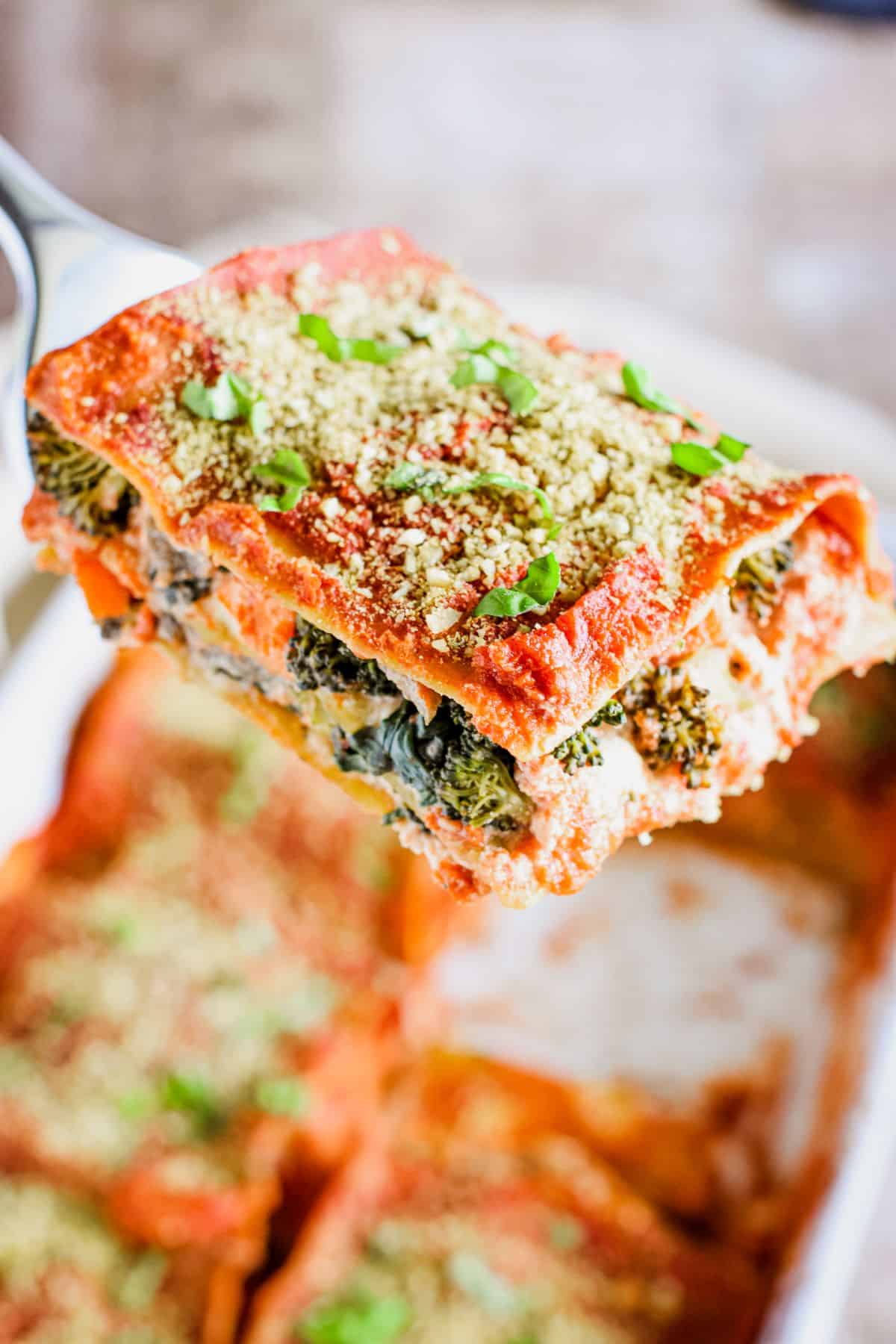 A metal spatula holding up a slice of vegan vegetable lasagna over a baking dish with the rest.