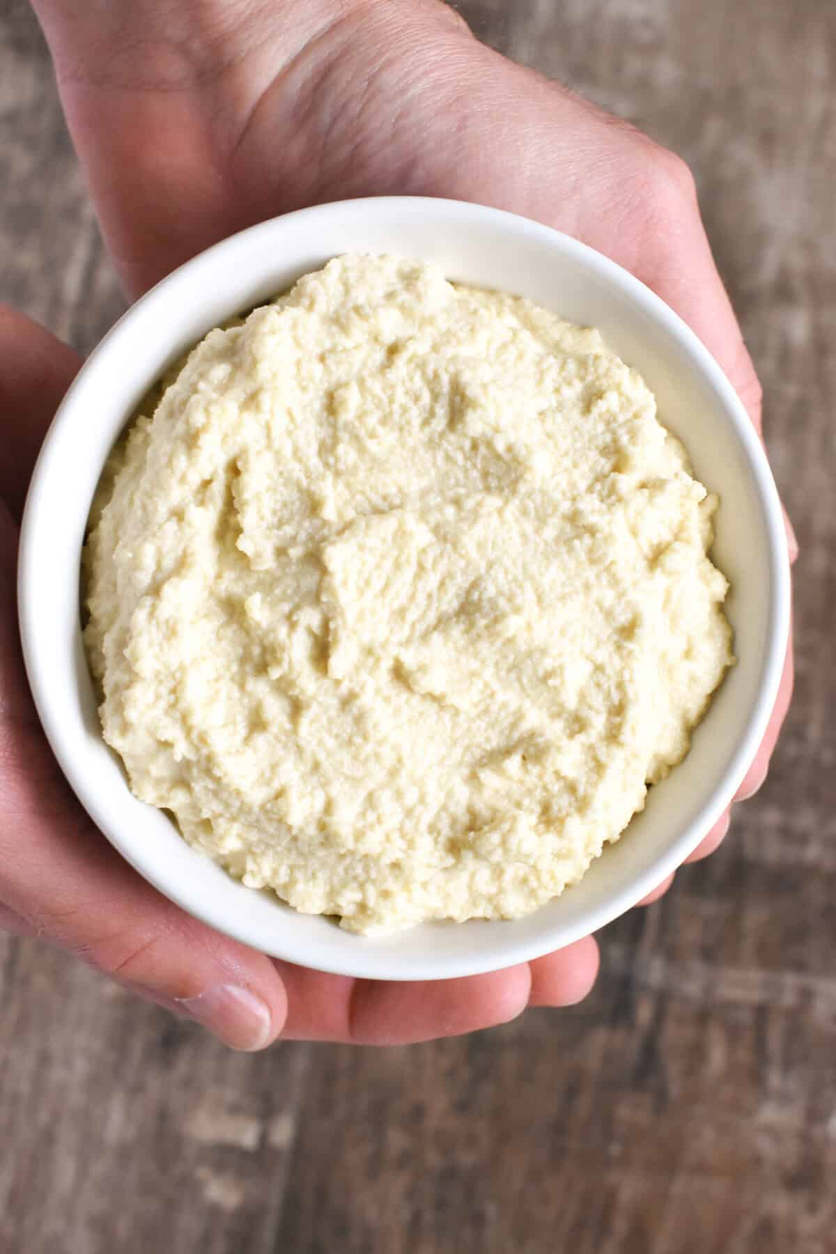 Almond ricotta in a white bowl with a spoon in it on a beige kitchen napkin.