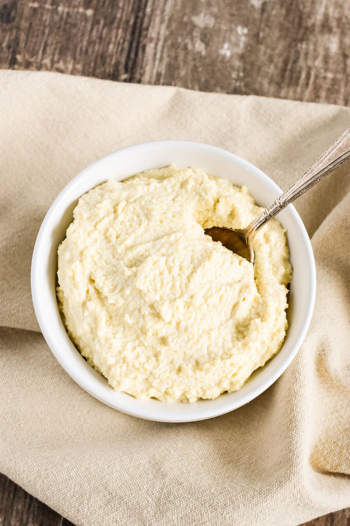 Almond ricotta in a white bowl with a spoon in it on a beige kitchen napkin.