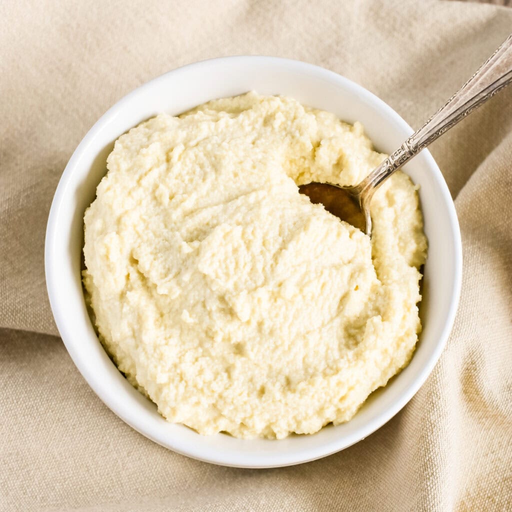 A white bowl with almond ricotta and a spoon in it sitting on a beige kitchen napkin.