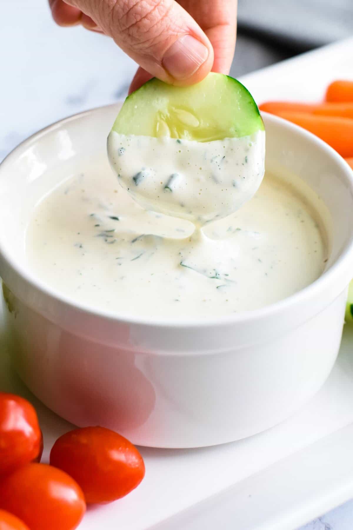 A hand dipping a cucumber slice into a bowl of cashew ranch dressing on a veggie platter.