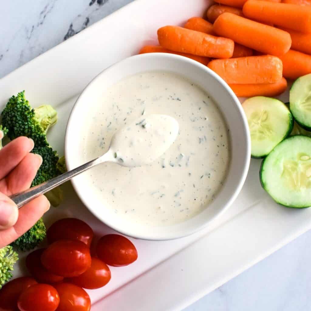 A person holding up a spoon with dressing on it over a bowl with the rest of it on a veggie platter.