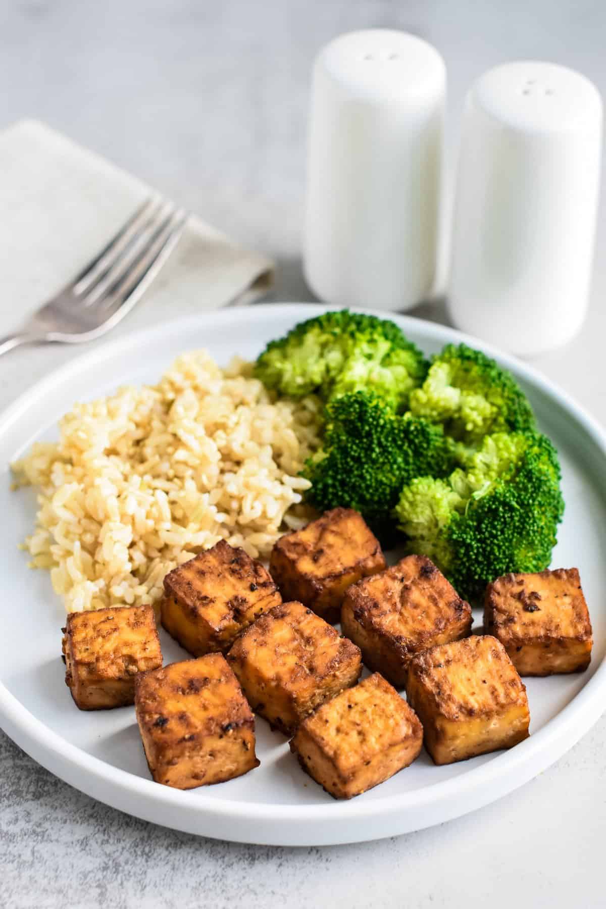 Baked tofu cubes on a white plate with brown rice and steamed broccoli.