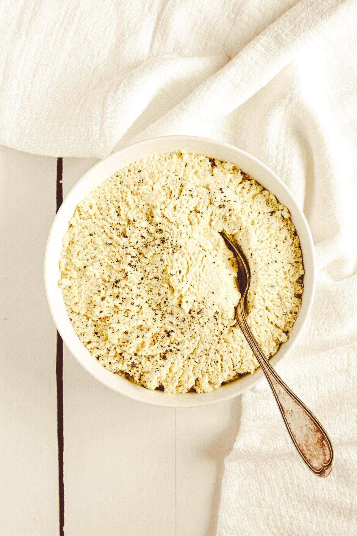 A white bowl of tofu ricotta sprinkled with black pepper and a spoon in the bowl.