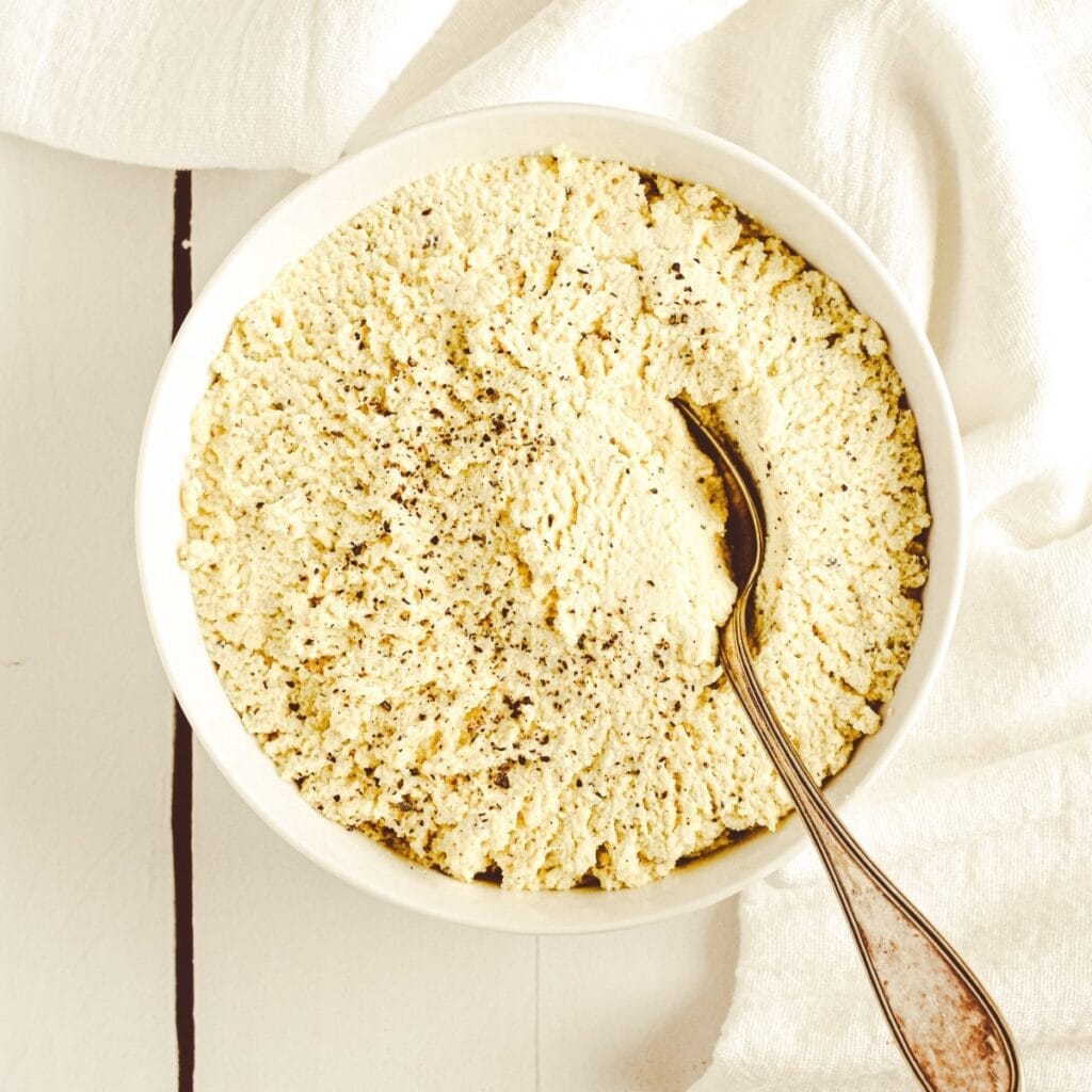 A white bowl filled with tofu ricotta with a spoon in it sitting on a white surface.