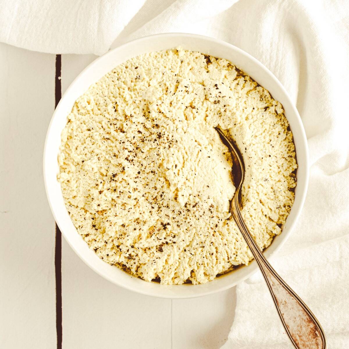 A white bowl filled with tofu ricotta with a spoon in it sitting on a white surface.